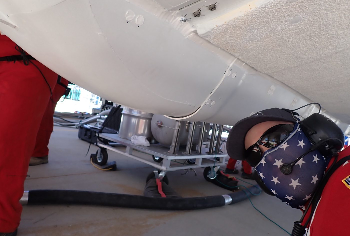 Ben Henderson in red coveralls, headset and face covering, crouched beneath the white underside of a rocket